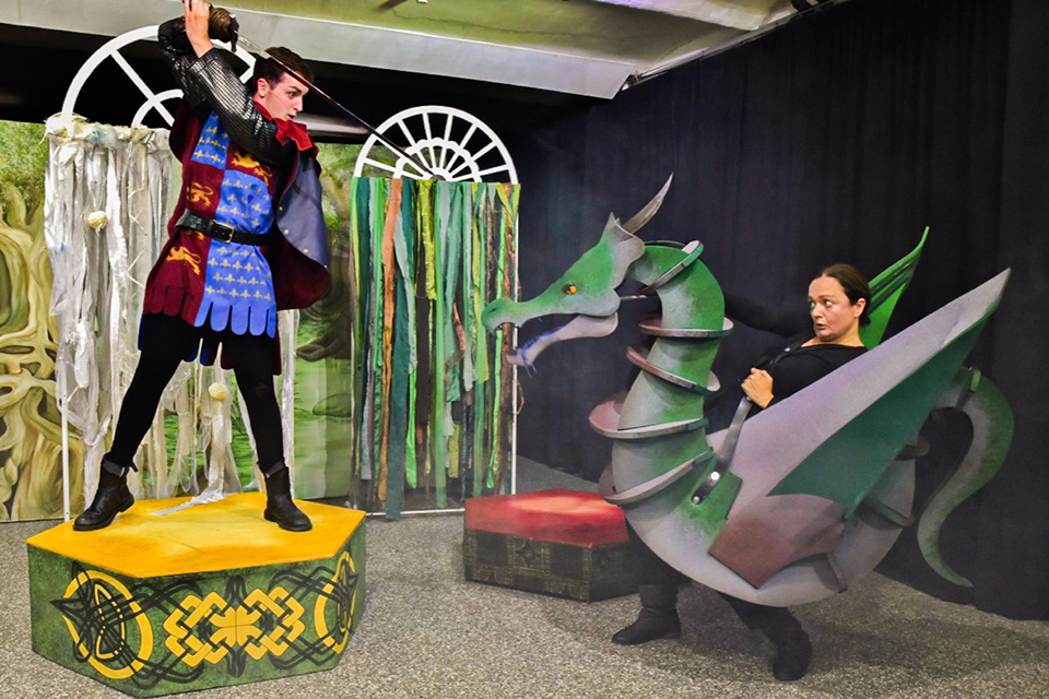 演劇The Green Knightの１シーンです。台の上に立った剣士役の男性が剣を握り、画面右側にいるドラゴン役の女性に向けて構えています。